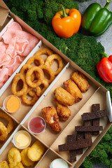 hot appetizers, beard toast, squid rings, spicy chicken wings deep-fried fries with soy sauce in cardboard packaging box against the background of green moss and red and yellow peppers