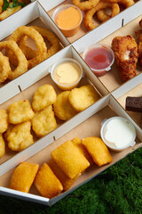 hot appetizers, beard toast, squid rings, spicy chicken wings deep-fried fries with soy sauce in cardboard packaging box against the background of green moss and red and yellow peppers