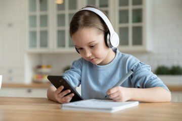Close up focused little girl wearing headphones using phone, studying online, sitting at table in kitchen at home, serious child holding smartphone, watching webinar, writing notes, homeschooling