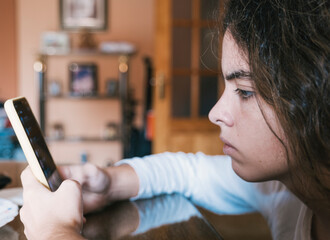 Teenage woman in pajama clothes sitting on the couch and with Smartphone in hand consulting ss...