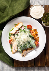 Portion of caesar salad with turkey roasted breast meat, cherry tomatoes, shredded parmesan cheese, salad dressing, salad leaves on a white plate on a wooden plank, green linen napkin.