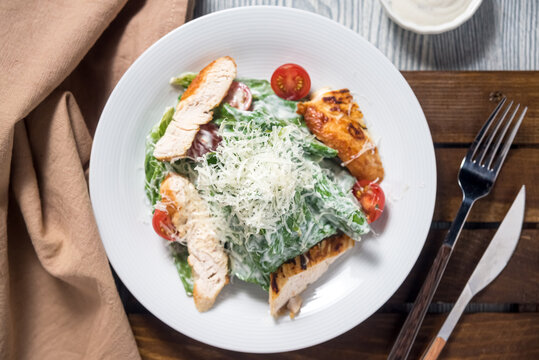 Portion of caesar salad with turkey roasted breast meat, cherry tomatoes, shredded parmesan cheese, salad dressing, salad leaves on a white plate on a wooden plank, green linen napkin.