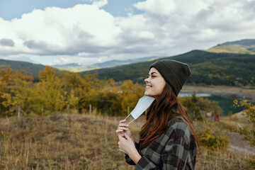 Happy woman with a medical mask and in a dark hat on nature in the mountains in the forest