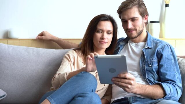 Happy young couple sitting on sofa, using tablet computer, browsing, laughing at funny photos, videos, having fun together, enjoying their weekend at home, Caucasian