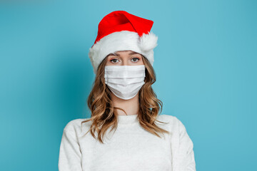 Young woman wearing protective medical face mask and santa hat stands isolated on blue studio background