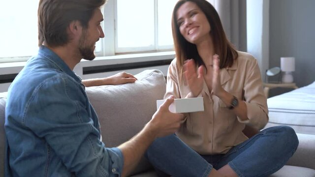 Young Caucasian couple happy lifestyle in home, handsome man holding gift box and make surprise to woman sitting on sofa. Husband give birthday or anniversary present for lovely wife