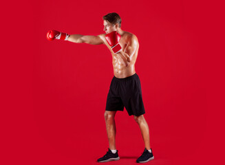 Side view of professional young boxer in gloves throwing punch on red studio background