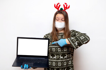 Concept of Christmas shopping, congratulations in pandemic. Young woman in medical mask, gloves and traditional red Christmas deer horns points finger at screen laptop. place for text
