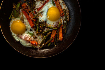 Two fried eggs in a pan with large slices of carrots, bell pepper and asparagus with spices and sauce. Close.