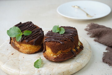 A picture focus on two chocolate cheese cake topped with chocolate glaze and mint leaves for garnish in marble board and grainy background