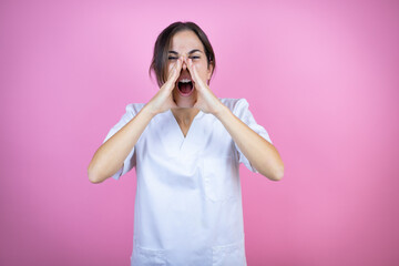 Young brunette doctor girl wearing nurse or surgeon uniform over isolated pink background shouting and screaming loud to side with hands on mouth