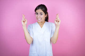 Young brunette doctor girl wearing nurse or surgeon uniform over isolated pink background gesturing finger crossed smiling with hope and looking side