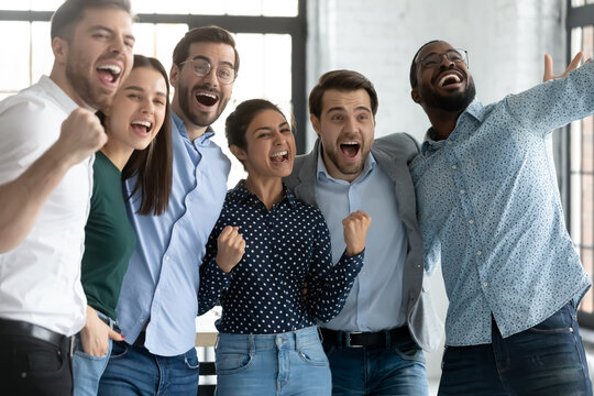 Yes. Overjoyed Friendly Diverse Millennial Coworkers Standing In Office Embracing Yelling Supporting Teammate Taking Part In Corporate Competition, Watching Favorite Sport Crew Triumph On Wide Screen