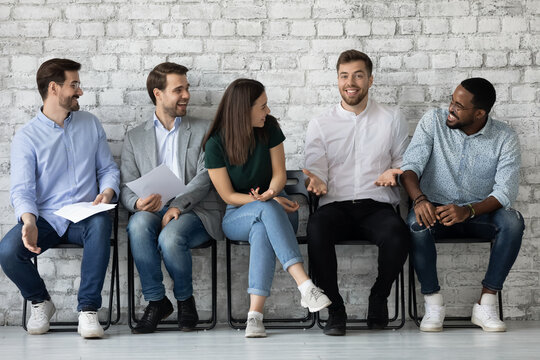 Diverse young people job applicants colleagues students sitting in row by wall waiting for interview conference exam being in good mood laughing resting relaxing talking having fun enjoying cute jokes