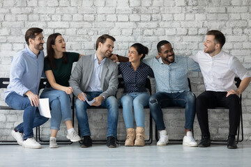 Friendly motivated company of diverse multiethnic colleagues students friends sitting on row of chairs hugging smiling having pleasant talk discussing affairs having fun spending good time together