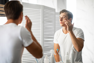 Handsome guy looking in the mirror applying cream on face