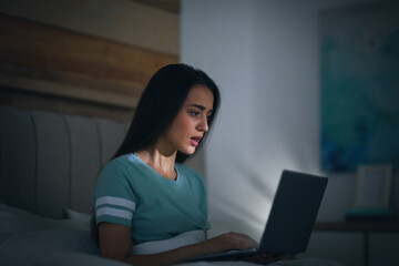 Fototapeta premium Emotional young woman with nomophobia using laptop in bed at night. Insomnia concept