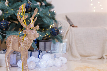 Golden shiny reindeer toy with white baubles, wrapped presents and decorated fir-tree, sofa on blurry background. Winter holidays, magic, Merry Christmas, Happy New Year concept. Copy space.