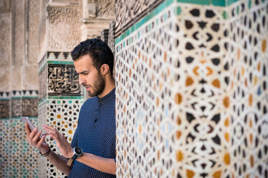 Young Muslim man using his mobile phone in casual clothing beside the wall with Moroccan decoration