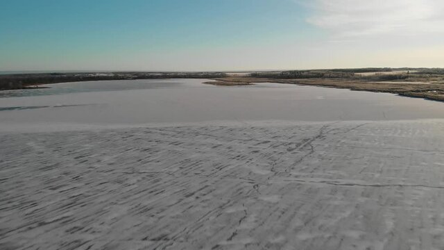 Drone flying high above a frozen lake that is partially covered by snow drifts.  Frost covered trees line the lakeshore and the sky is blue with thin white clouds.
