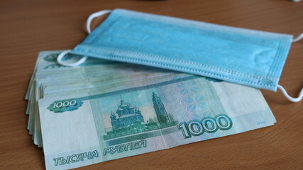 a pack of Russian bills of one thousand rubles scattered on the surface of the table and a clean blue medical mask symbolizing individual protection from the virus