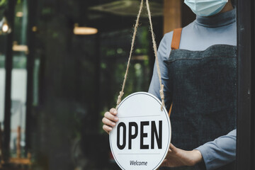 Reopen. friendly waitress wearing protection face mask turning open sign board on glass door in...