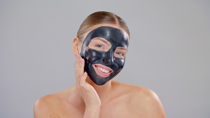 Close-Up Beauty Portrait Of Young Caucasian Blonde Woman With Blue Eyes She Applying Black Mask On Face And Smiling On Gray Background She Using Hand And Finger On Healthy Skin Care Concept