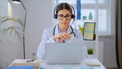 Young Female Doctor Wears White Coat, Video Calling Distant Patient on Laptop. Doctor Talking to...