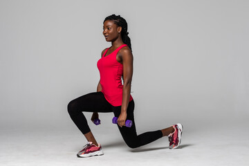 Attractive young african american sports fitness woman in sportswear posing working out isolated on gray background. Sport exercises healthy lifestyle concept. Make exercise doing lunges with dumbbell