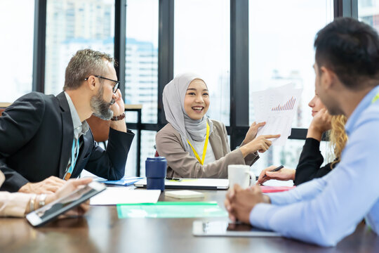 Asian Muslim Business Woman In Hijab Headscarf Sitting On Wheelchair Presenting Of Her Work To Corporate Colleagues In Meeting In The Modern Office. Diverse Corporate Colleagues And Multicultural