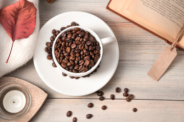 A white Cup full of coffee beans on a light beige wooden background with a book and a sweater. Top view with space to copy.