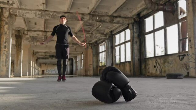Boxer jumping using jump rope while warming up in abandoned factory in slow motion. Boxing gloves at foreground. Young boxer skipping. Sport concept. Work out of mma fighter. Full length in 4K, UHD