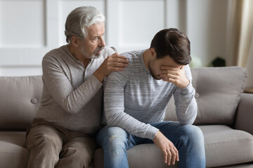 Caring loving mature father calming hugging upset adult son, sitting on couch, senior older...