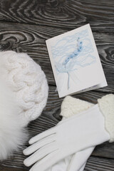 Winter knitted hat with fur and gloves. White. Next to the greeting card. They lie on black pine boards.