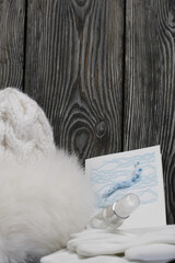 Winter knitted hat with fur and gloves. White. Nearby is a greeting card, a medical mask and an antiseptic. They lie on black pine boards.