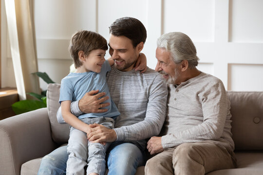 Happy three generations of men hugging, sitting on cozy couch at home, cute adorable little boy cuddling with young father and mature grandfather on sofa, family spending leisure time together