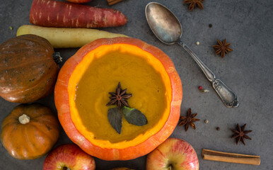Squash soup in pumpkin bowl. Top view photo of pumpkin, apples, carrots anise stars and cinnamon sticks. Grey background. Autumn food close up photo. 
