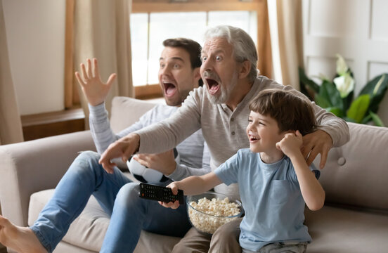 Overjoyed little boy with father and grandfather celebrating goal, sitting on couch with popcorn and soccer ball, excited happy three generations of men watching tv, supporting favorite football team