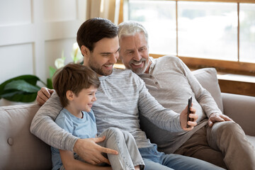 Obraz premium Close up happy three generations of men using smartphone together, having fun with gadget, overjoyed little boy with mature grandfather and father looking at phone screen, sitting on couch at home