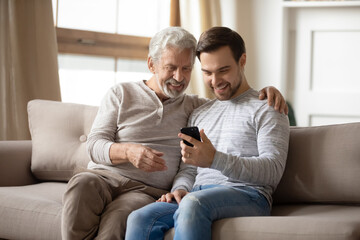 Happy older father and adult son hugging, using phone at home together, smiling mature grandfather...