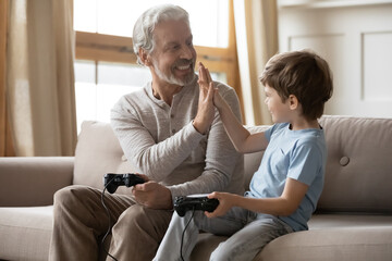 Overjoyed mature grandfather and adorable grandson giving high five, playing video game, celebrating win, excited older man and little grandchild boy having fun, holding controllers, sitting on couch