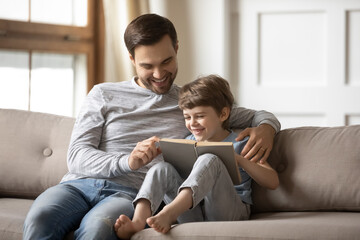 Loving happy father teaching adorable son to read, smiling dad and little boy child hugging, sitting on couch, holding book with fairy tale story, family spending weekend together, leisure time