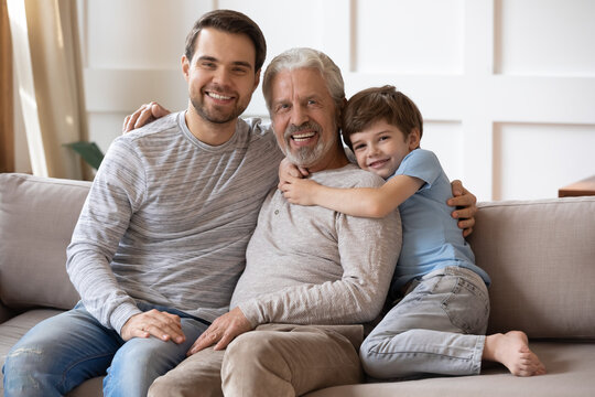 Family portrait smiling mature grandfather, young father and little boy hugging, sitting on couch at home, happy three generations of men looking at camera, posing for photo in living room