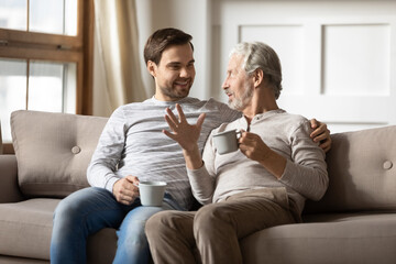 Happy mature father and adult son drinking tea together, chatting, enjoying pleasant conversation, leisure time, sitting on cozy couch at home, smiling grandfather and grandson holding cups