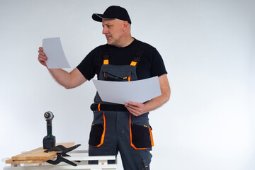 Carpenter in a gray jumpsuit and a baseball cap. Joiner examines the drawings. A man stands in a carpenter shop. The concept of woodworking. Manufacturing of furniture to order.