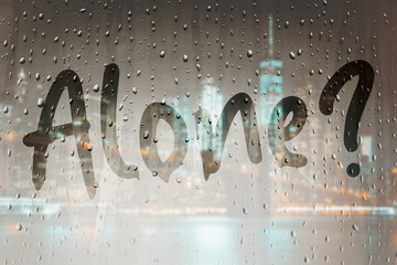 Autumn rains and wet window. The inscription on the sweaty glass - Alone. Depression and lonely in big city concept. City lights blurred background.