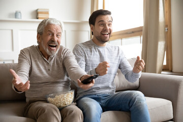 Overjoyed mature father with son watching tv, soccer match, having fun together, sitting on cozy...