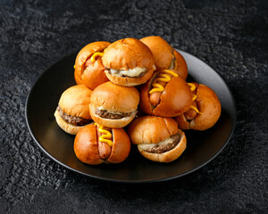 Mini hamburgers, burger and hot dogs with yellow mustard on black plate