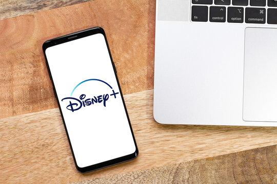 Disney + Logo, Disney plus symbol on a smartphone screen with labtop and wooden desk background, top view