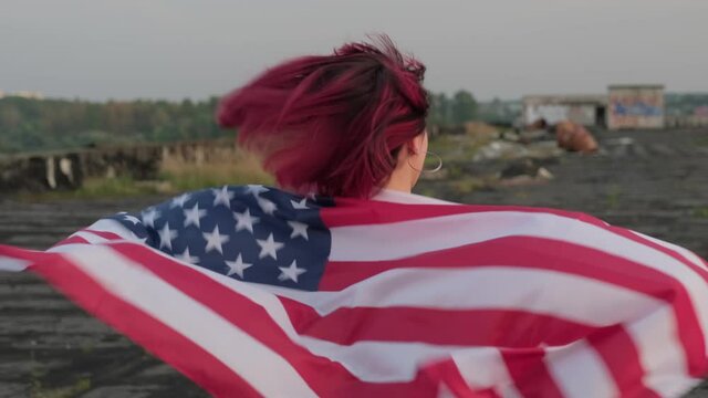 Happy woman with USA national flag running outdoors at sunset. US Independence Day. Female with American flag on her back. Tracking shot in 4K, UHD